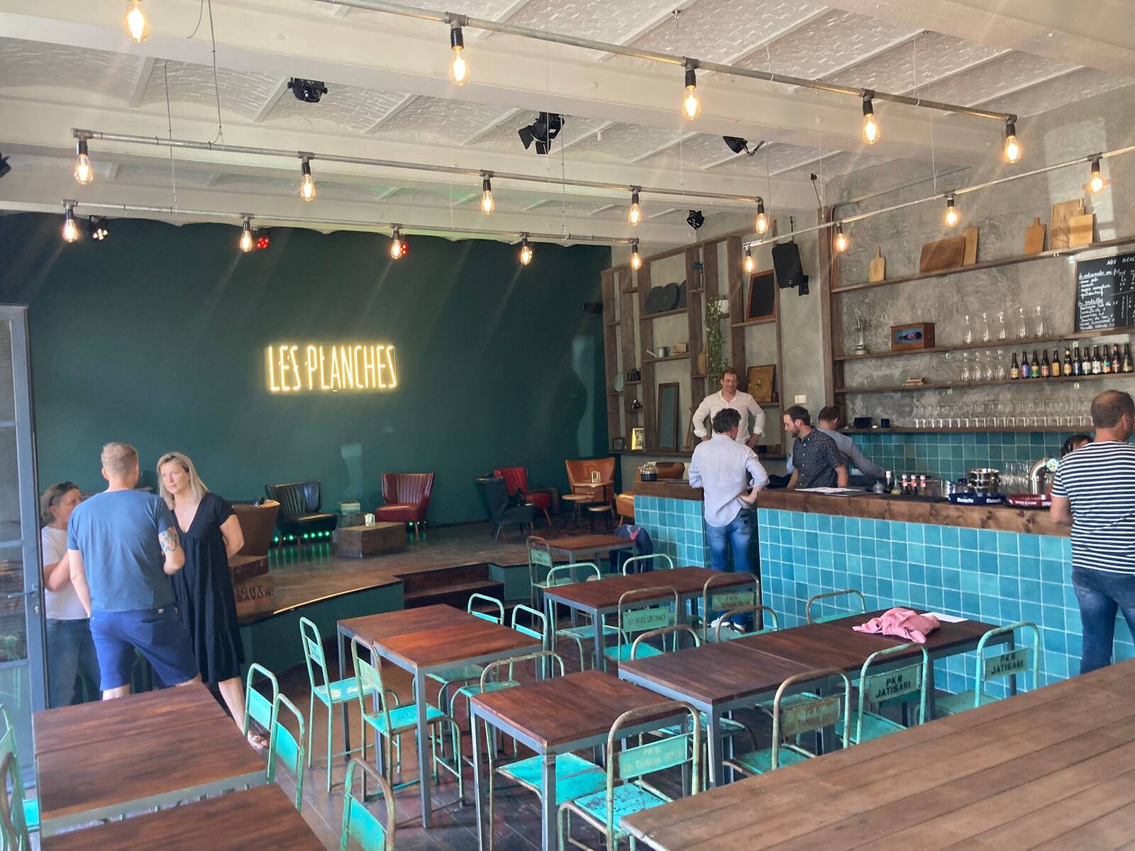 La grande salle avec ses tables en bois, le bar carrelé turquoise et la scène au fond